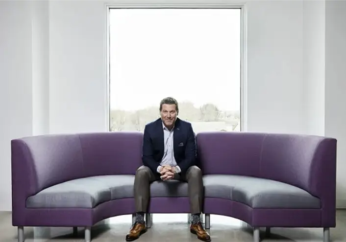 Man sitting happily on a curved purple sofa with a large window behind him revealing a lot of natural light
